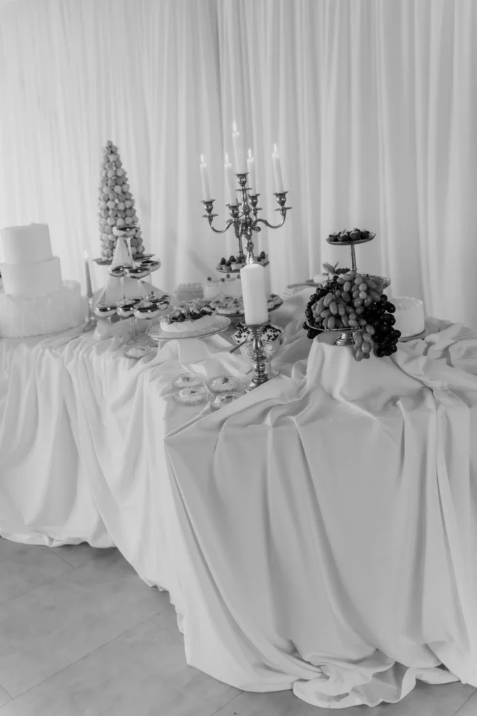 Elegant black and white photography of a lavishly decorated dessert and buffet table by Catering Majetic, featuring candelabras and tiered displays.